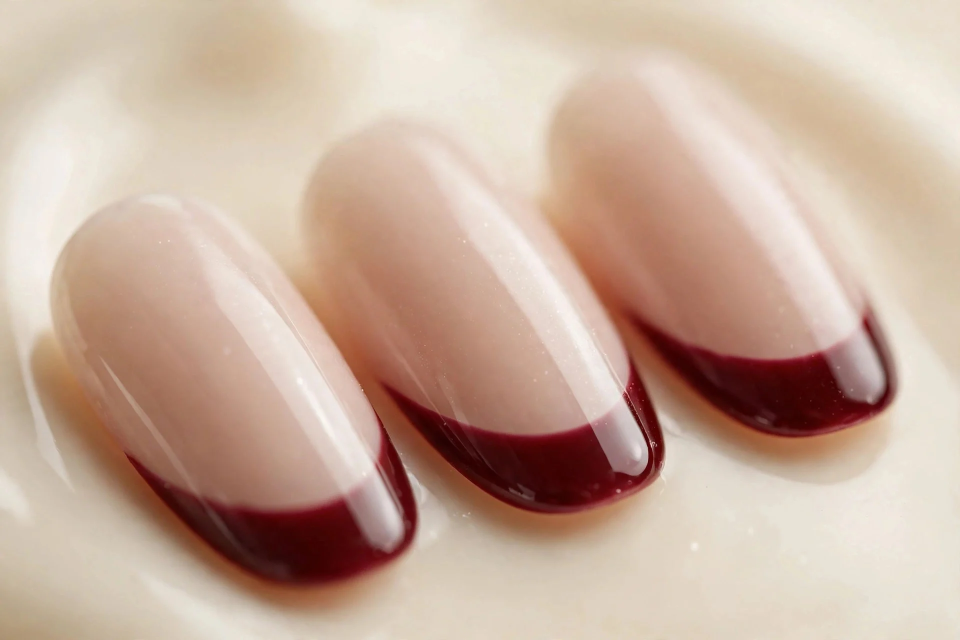 Almond French nails with cherry wine tips over sheer nude, glossy, cream background