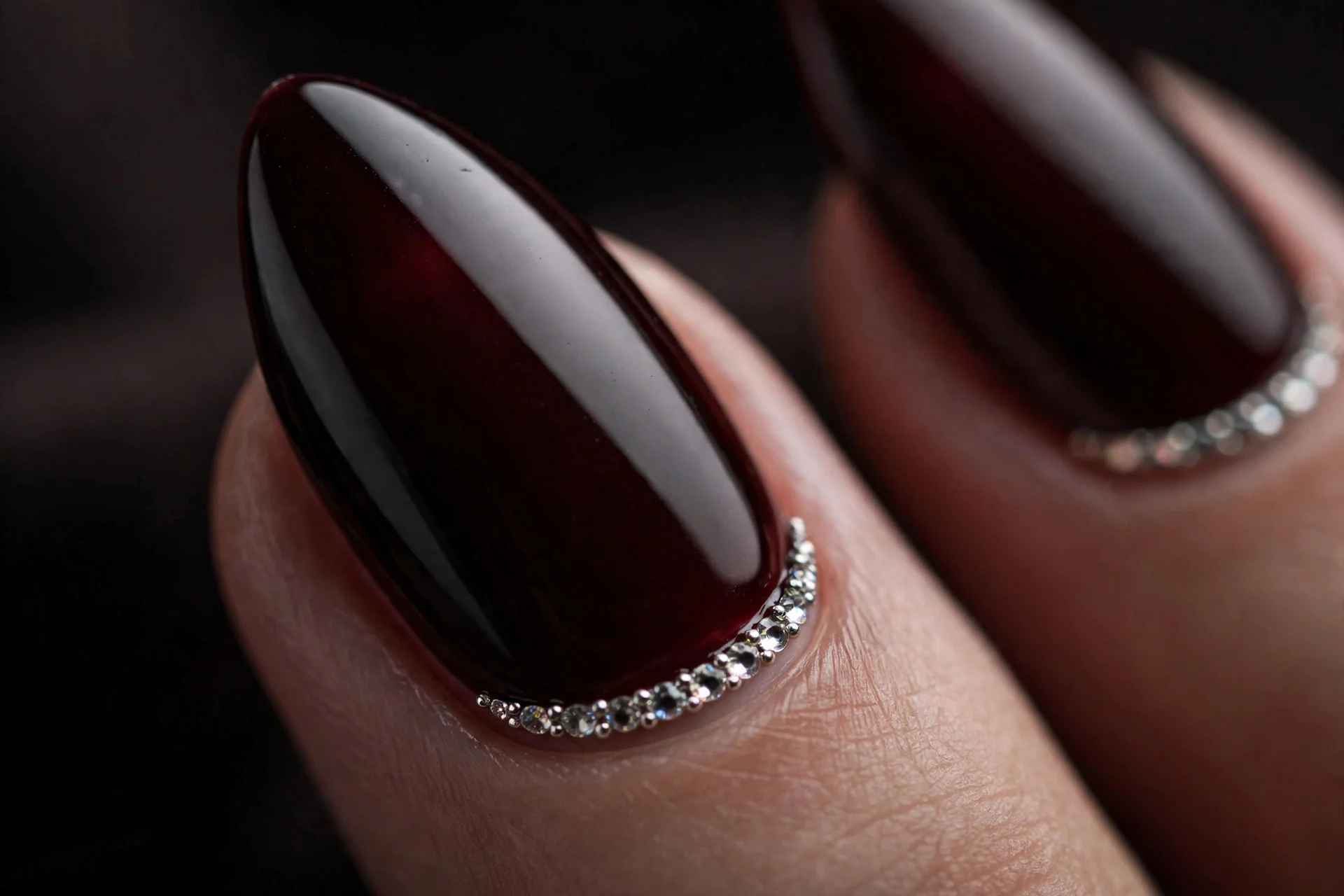 Almond nails in black cherry with tiny rhinestone lines along cuticles, glossy, dark background