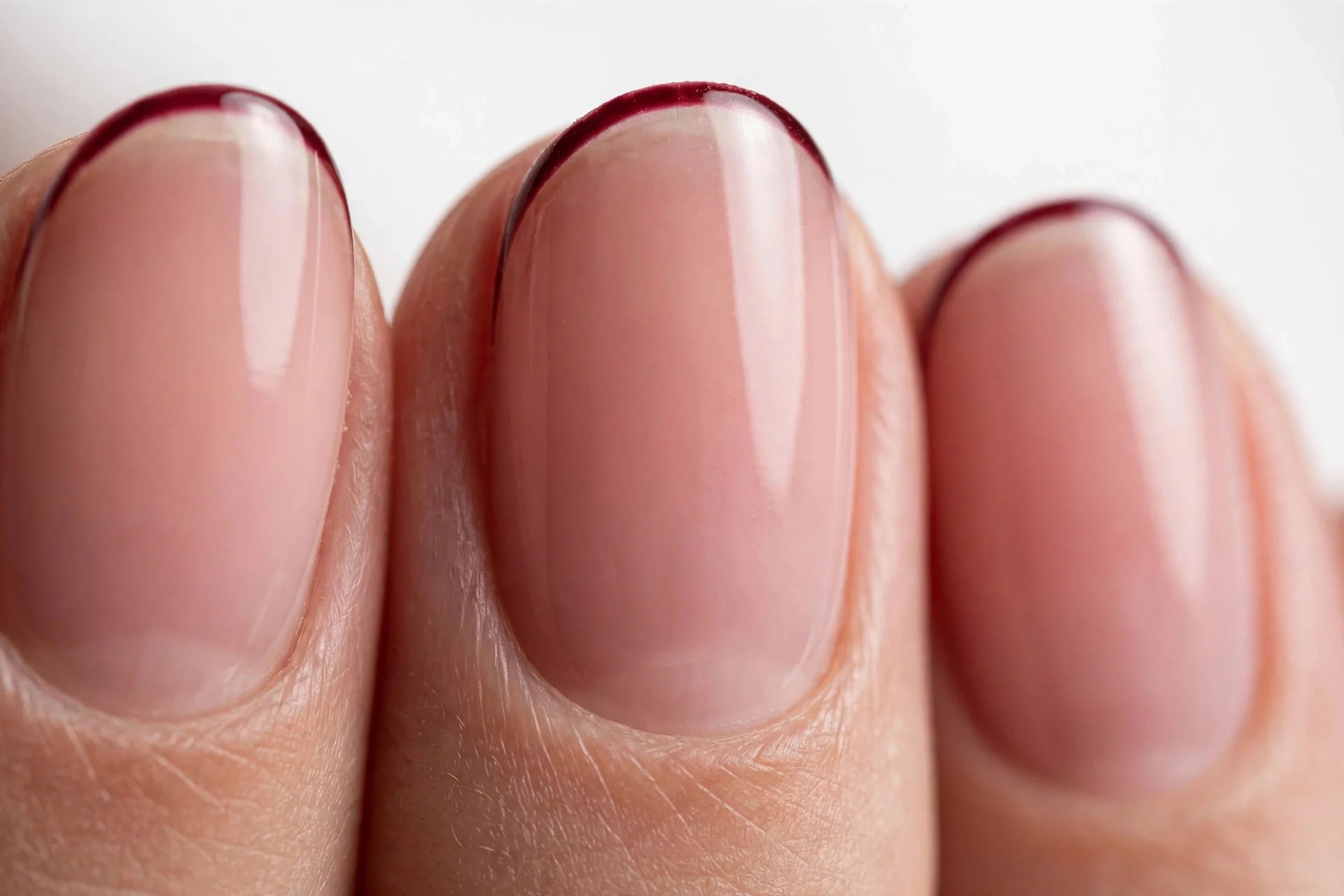 Oval French nails with thin cherry wine micro tips over sheer blush, glossy, white background