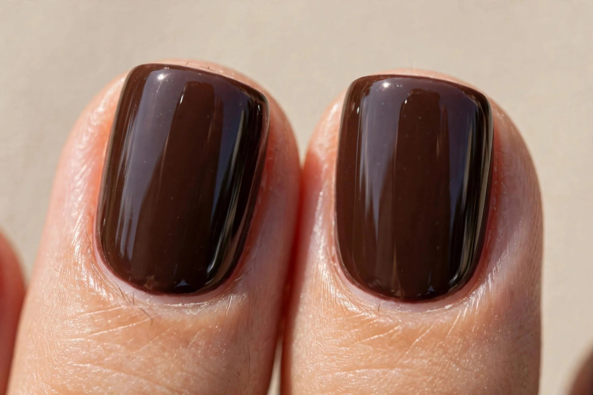 Close-up of two short square fingernails in deep glossy hot-fudge brown, mirror-like shine, neutral beige background