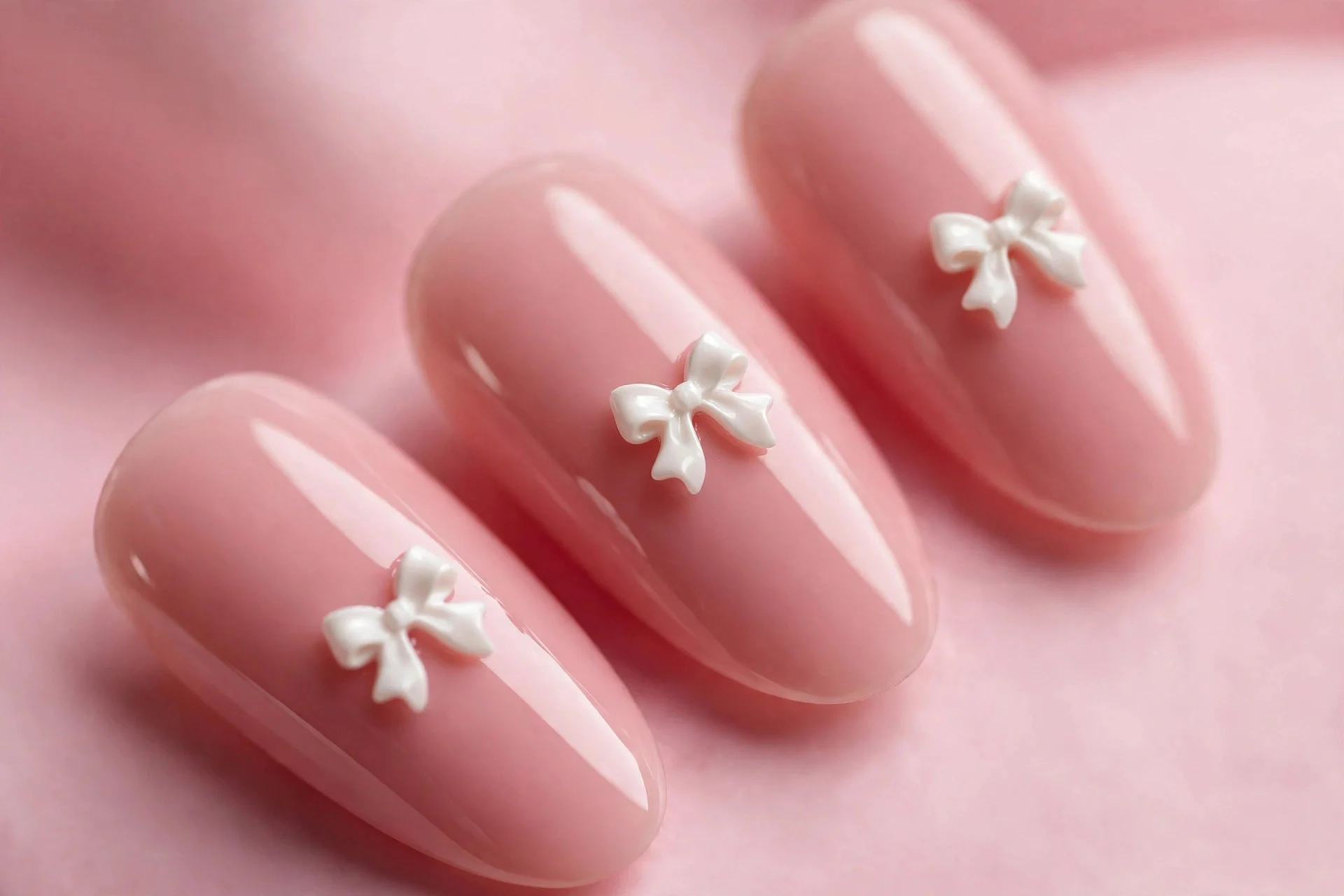 Almond blush pink nails with tiny white 3D bow charms, glossy, pink background
