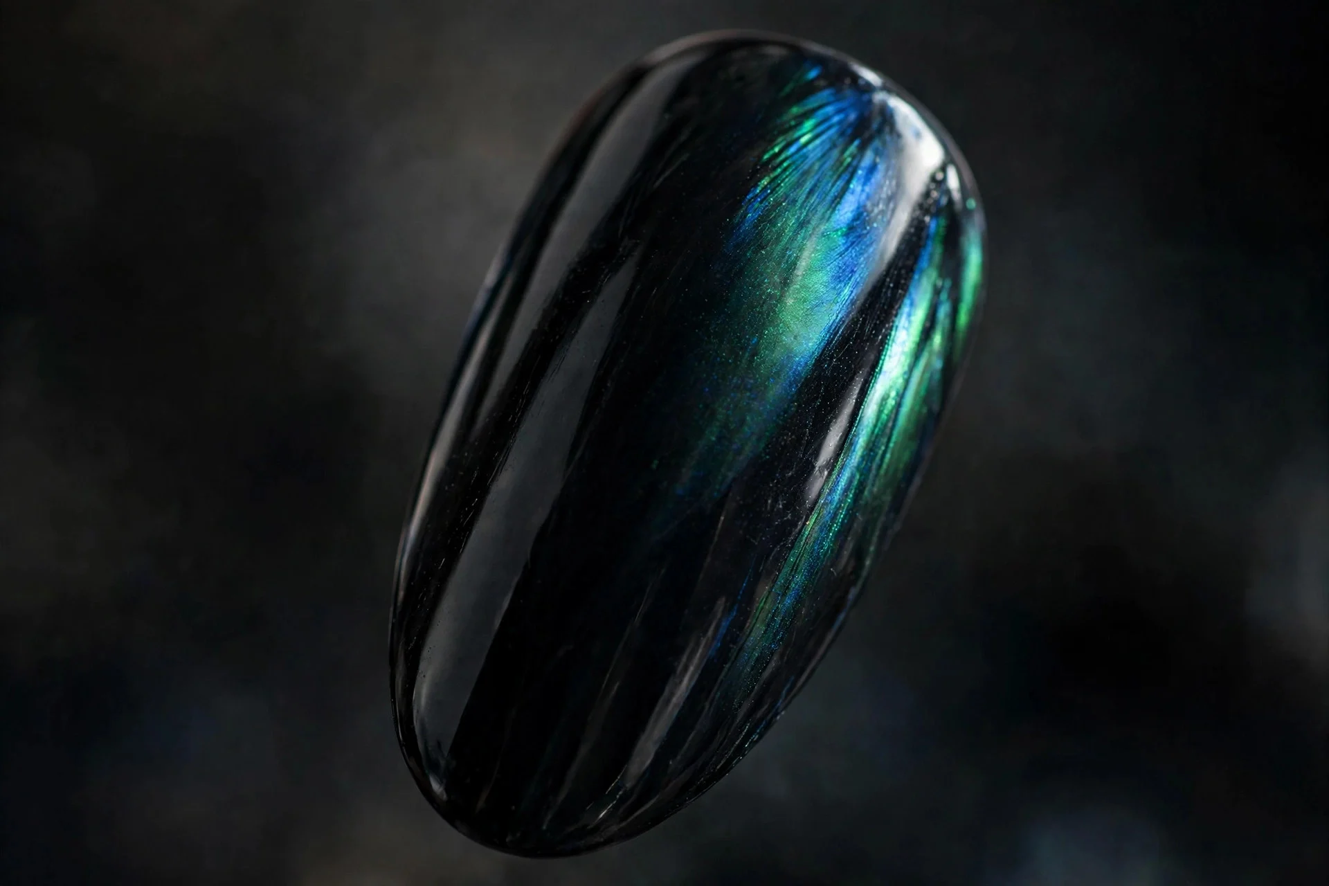 Almond fingernail in iridescent black chrome flashing blue-green duochrome like raven feather, dark background