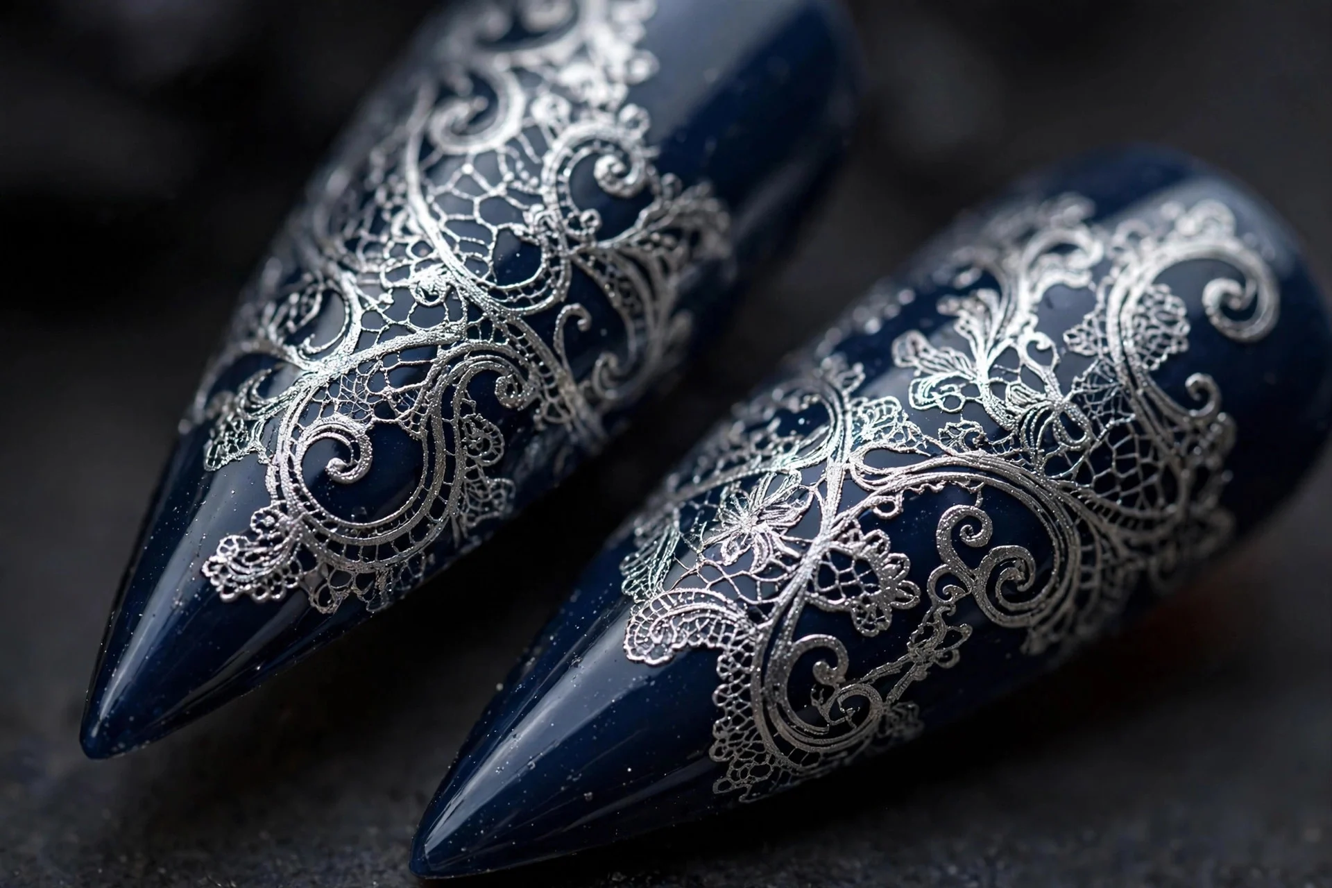 Stiletto nails with silver lace stamping over deep navy, intricate scrolls, glossy, dark background