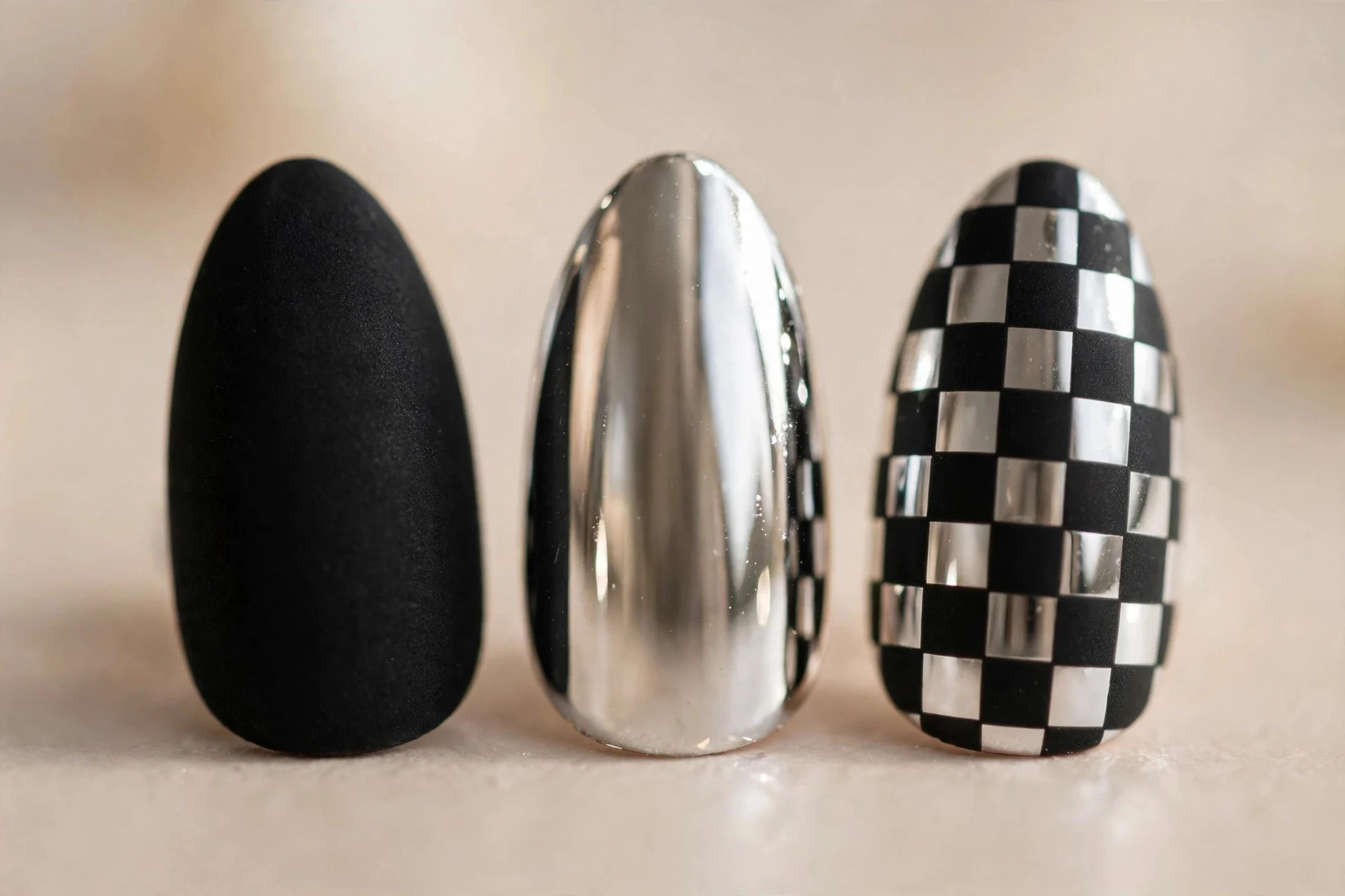 Extreme macro of three almond nails in matte black, mirror chrome and black chrome checkerboard, beige background