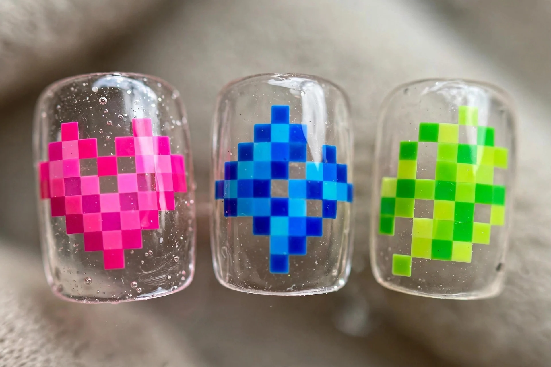 Extreme macro of three square nails with pixelated blocks in hot pink, blue and lime over clear base