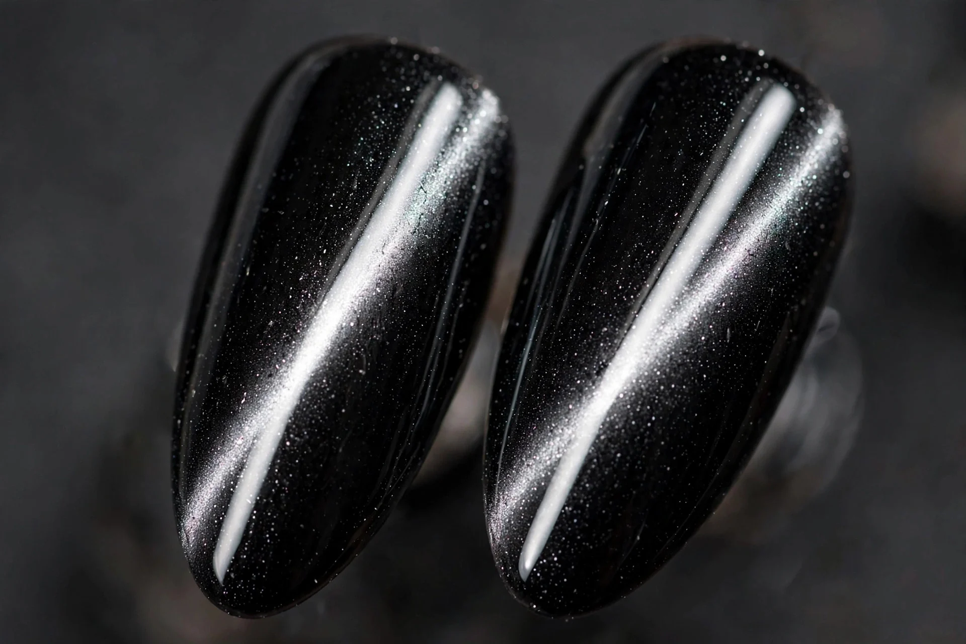 Extreme macro of two almond fingernails, black cat-eye gel, sharp silver light band, glossy, dark gray background