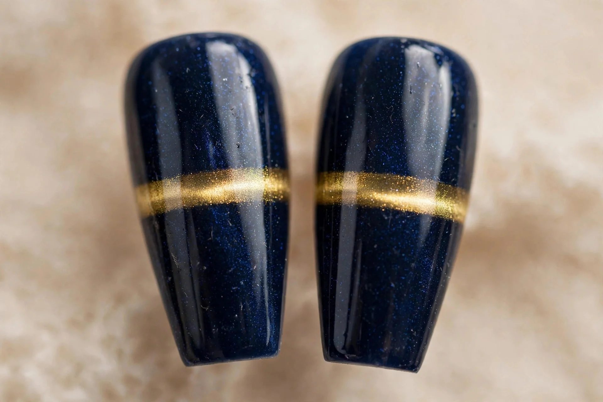 Extreme macro of two coffin fingernails, deep navy cat-eye gel, gold band, glossy, beige background