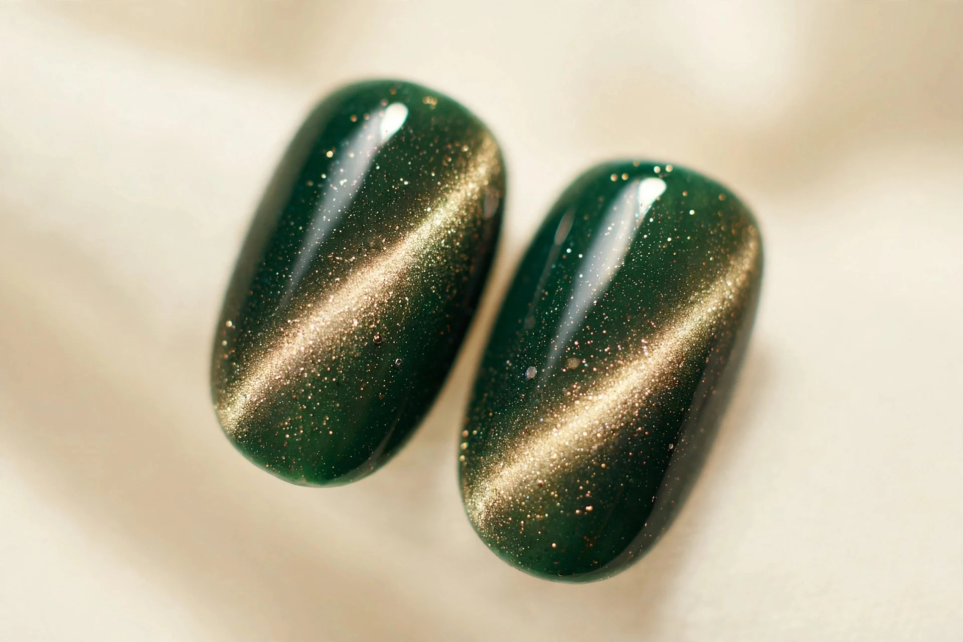 Extreme macro of two short oval fingernails, forest green cat-eye gel, champagne glow, glossy, cream background